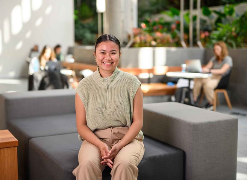 A smiling female student sits in front of a plant wall on campus.