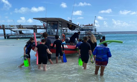 A group of students wade through knee deep ocean water to load science equipment onto a boat.