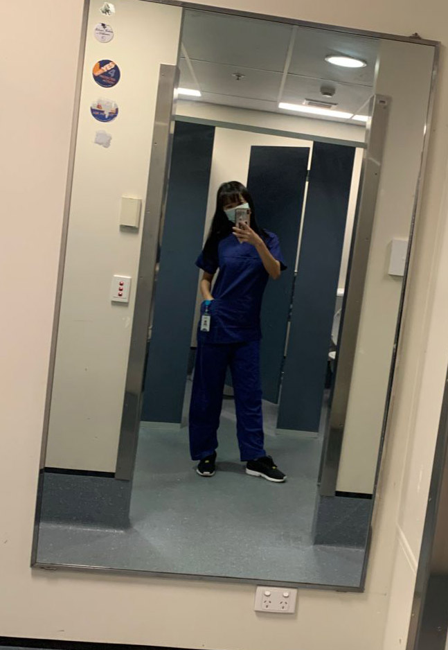 A woman in a staff change room photographs herself wearing surgical scrubs.