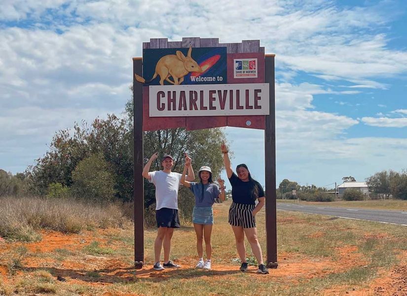 Three students stand under a Welcome to Charleville sign in the outback.