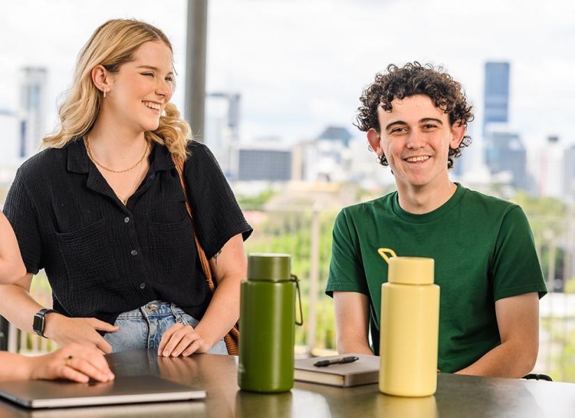 A smiling male student in a green t-shirt, Luke Gilbert, sits with a group of friends at a table on campus. In front of him is a note book, pen and water bottle.