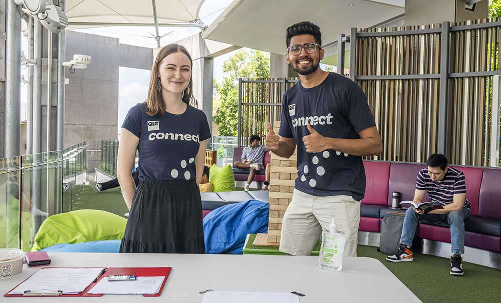 A male and female student in QUT Connect t-shirts take a break between chatting to students at an event. They are smiling at the camera, infront of them is a desk with a sign up sheet.