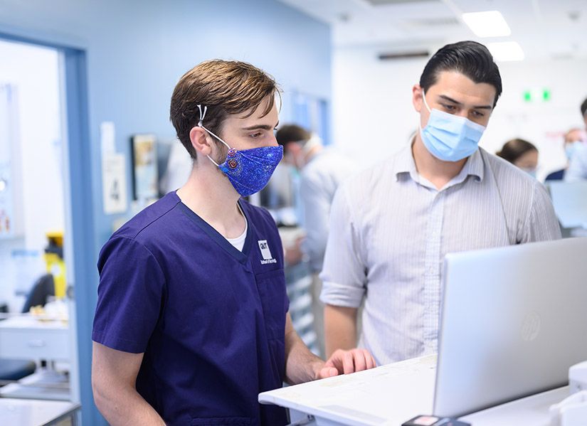 A male nursing student in scrubs and a mask stands in a hospital hallway. He is looking at a laptop while a supervisor explains a process.