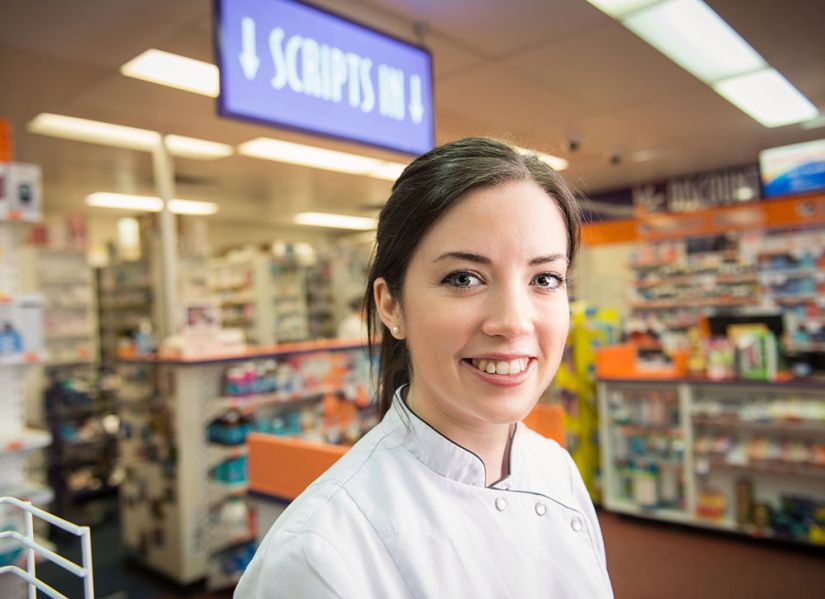 A smiling female pharmacy student stands in front of shelves in a community pharmacy.