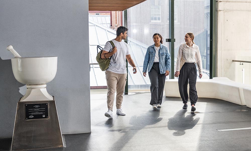 A male student with a backpack and two female students chat as they walk through a corridor.