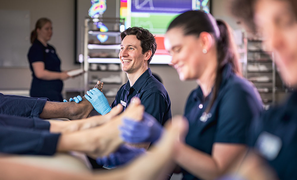 A row of podiatry students practice performing a foot exam on fellow students sitting on benches. Everyone is laughing because they feel silly at first before they become more proficient.