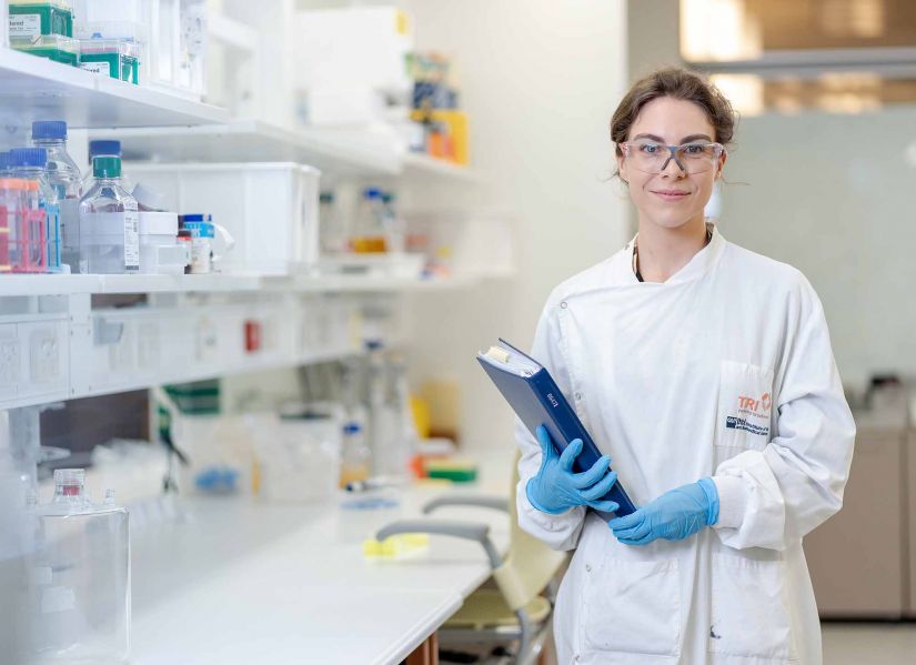 A woman wearing a labcoat, glasses and gloves stands next to a bench in a modern laboratory.
