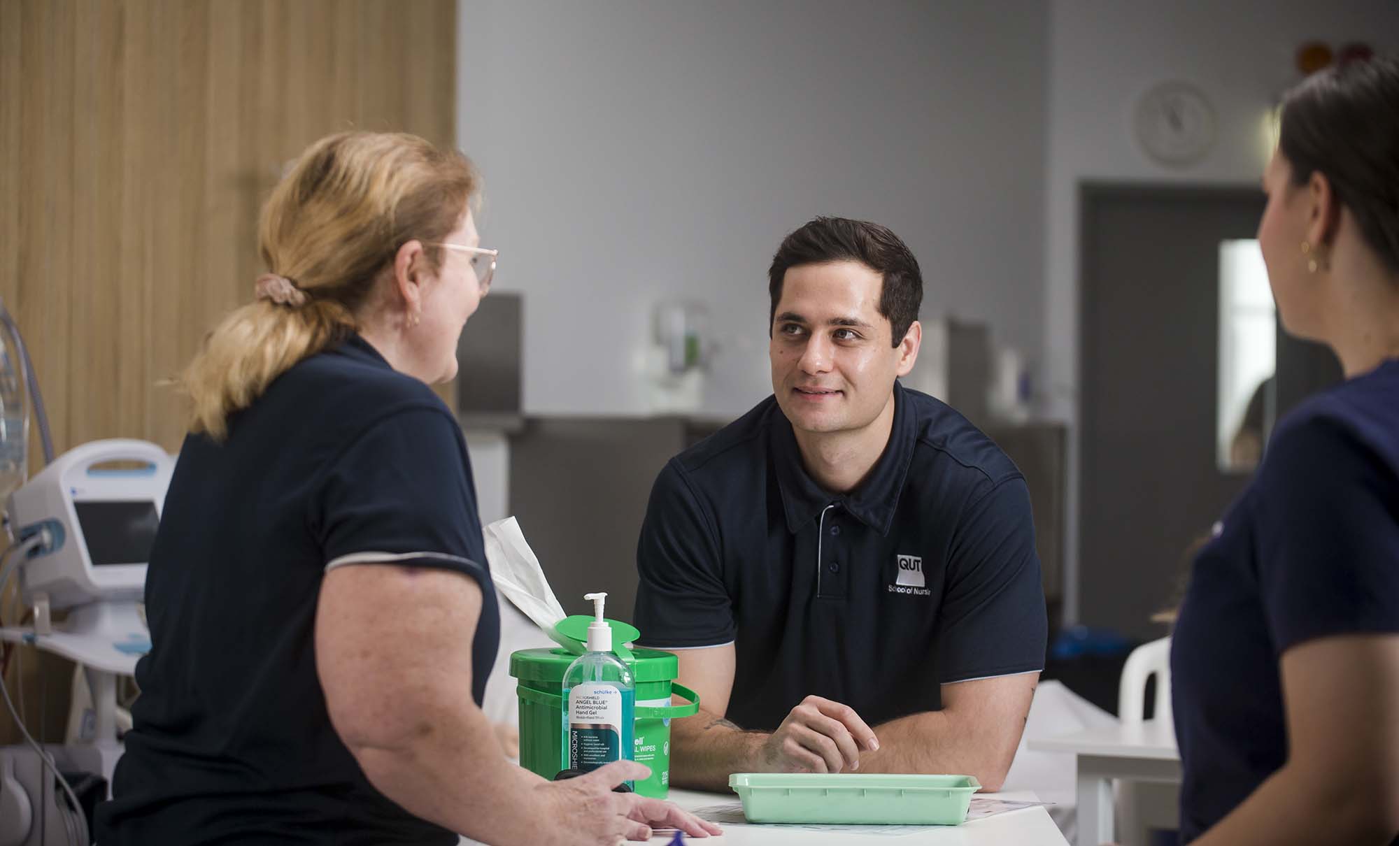 A male nurse leans of a bed in a classroom that looks like a hospital ward. He is smiling and looking up at a teacher who is talking to him.
