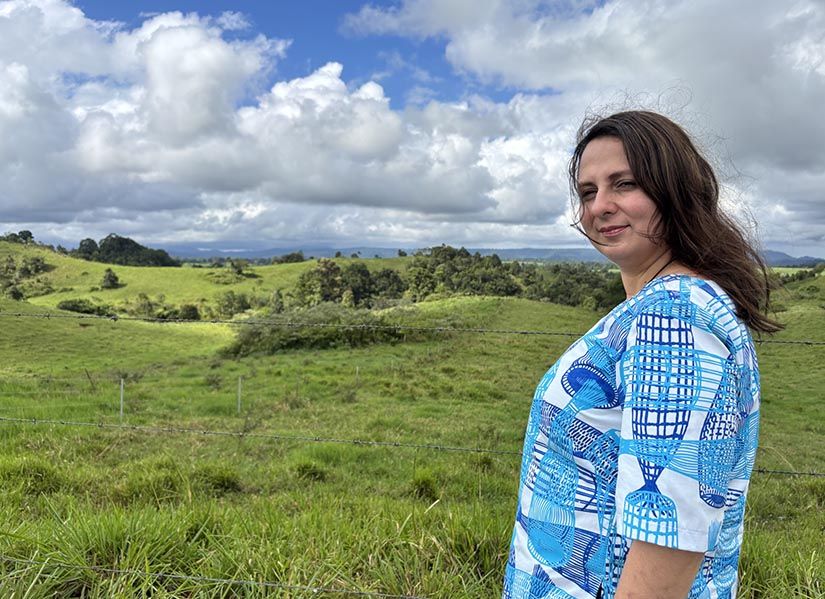 A woman wearing a shirt featuring Indigenous Australian artwork stands outside on a sunny, windswept hillside.