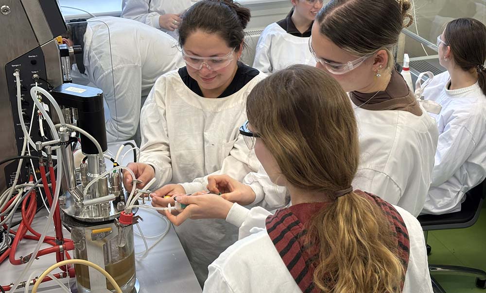 Three female students in lab coats manipulate a complicated container on the bench in front of them. It is processing brown liquid through an array of connected tubes.