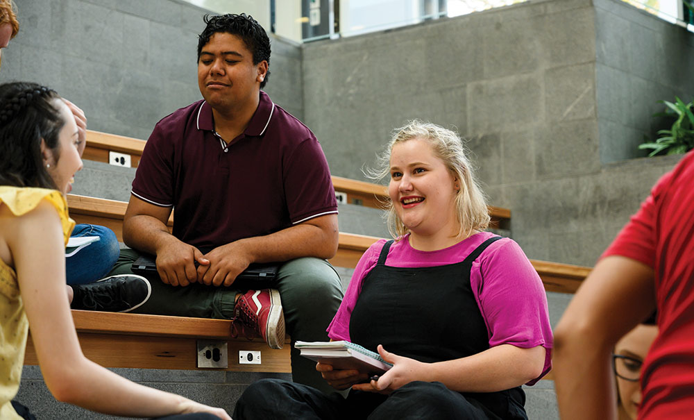 A female uni student in a larger body wearing navy overalls and a pink shirt smiles as she sits in a group listening to other students.