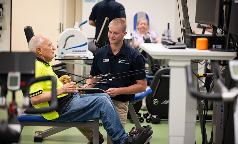 A male exercise physiologist watches as a patient performs an exercise on a rowing machine.