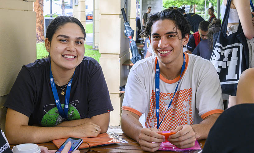 A male and a female student smile as they sit at a registration table for a uni event.