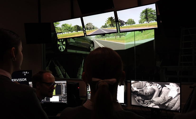 A researcher sits infront of monitors. He's watching a driver in a car on a mechanical rig navigate a driving a simluation.