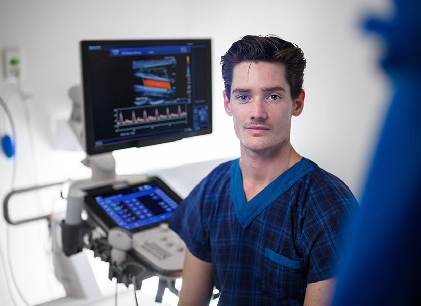 A man in scrubs sits in a clinical setting surrounded by sonography equipment.