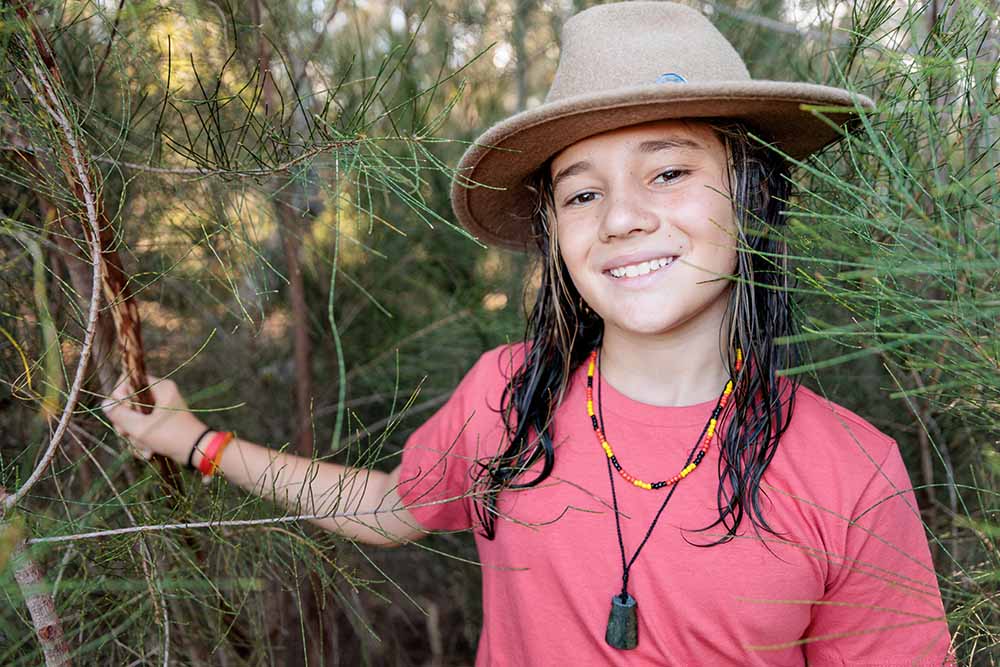 A young girl stands outdoors, she's wearing a broad-brimmed hat and a necklace with yellow, black and red beads.
