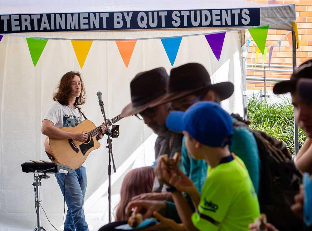 A QUT musician, playing a guitar and singing into a mircophone performs under brightly coloured banners at Open Day. In front of them a crowd listens while eating sausages.