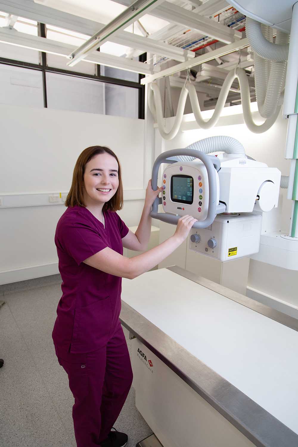 Zoe is wearing maroon scrubs and standing in front of an imaging machine, adjusting the settings for a treatment simulation.
