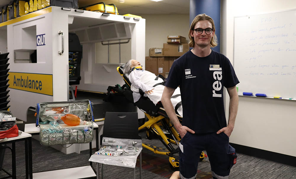 A paramedic science student stands in front of a life-sized ambulance simulator. Directly behind him is a patient mannequin on a gurney.