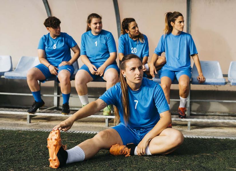 Four women in soccer uniforms sit on the bench and another player stretches on the ground in front of them. They are frowning as they watch what's happening on the field.