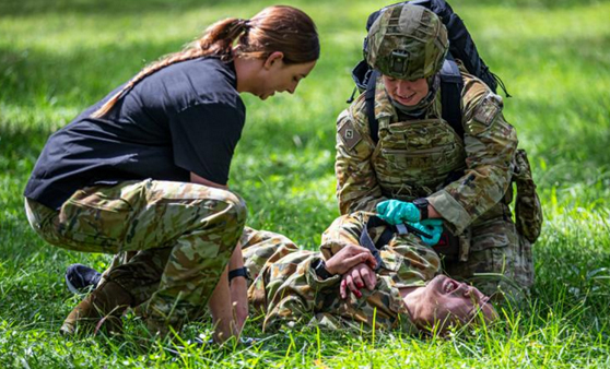 An army medic crouches down to treat a patient lying on the ground clutching their arm. Both patient and medic are watched by a supervisor crouching next to them.