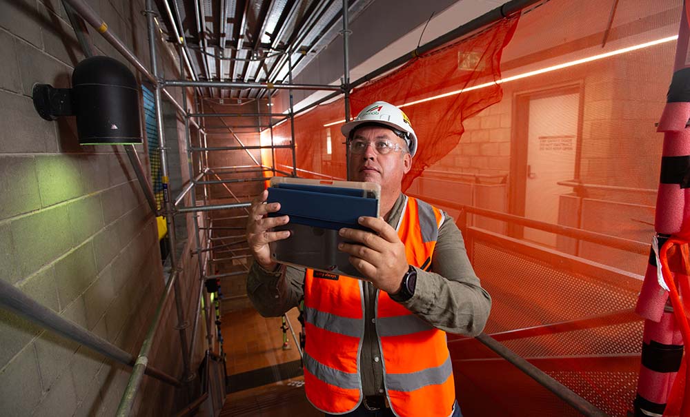 A man in a high visibility vest and a hard hat holds up an ipad while doing a safety audit on a construction site.