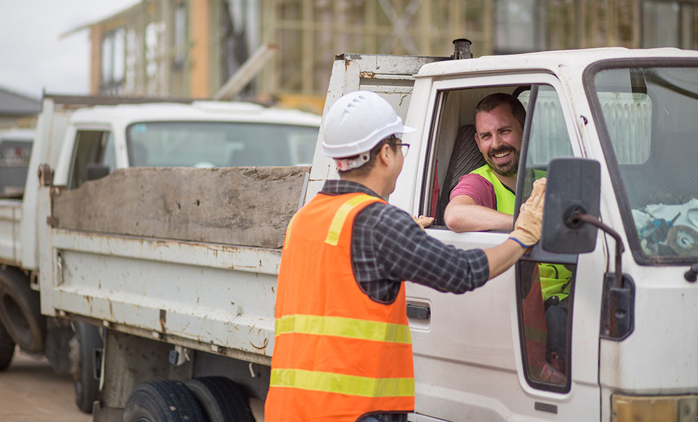 A tradie in a white ute arrives on a work site and chats to a man wearing a high visibility vest.