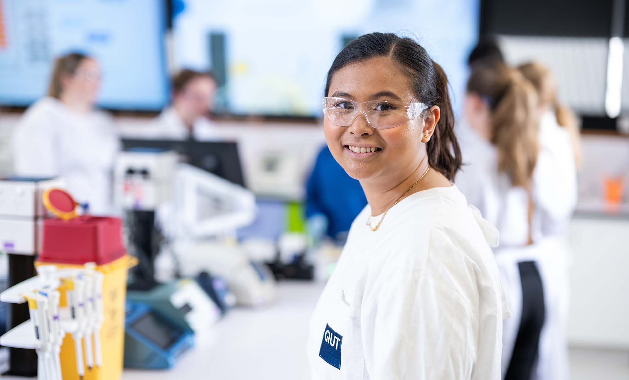 A smiling female student in a white gown and protective glasses stands in a laboratory.