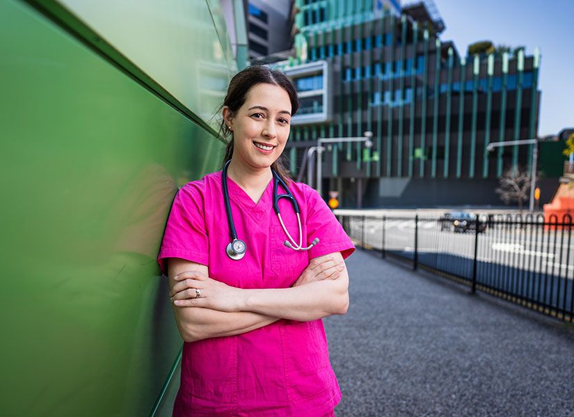 A smiling woman wearing pink scrubs stands outside a hospital building.