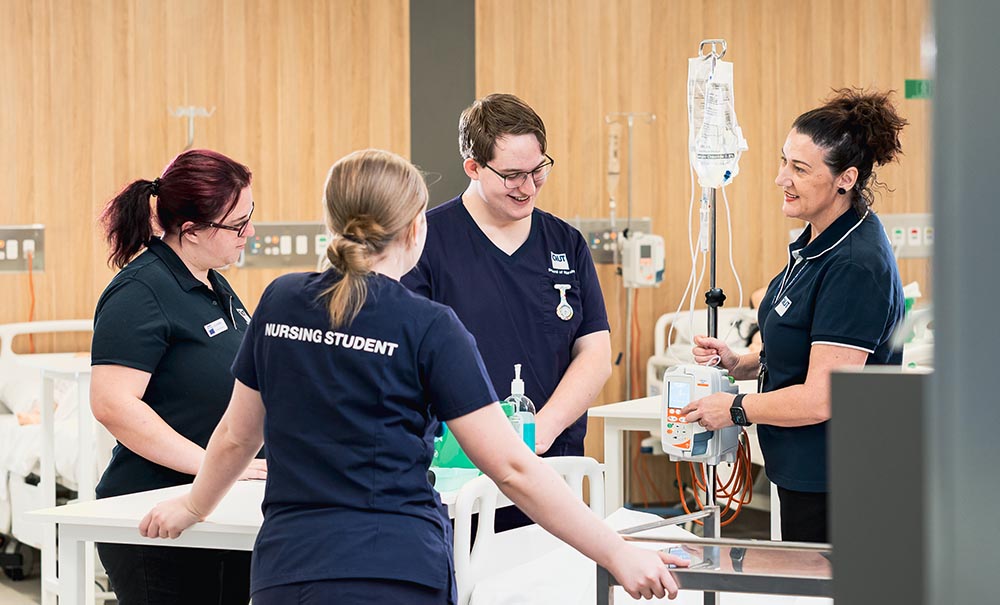 Three students in medical scrubs stand in front of a hospital bed listening to a teacher explain what they will be doing.