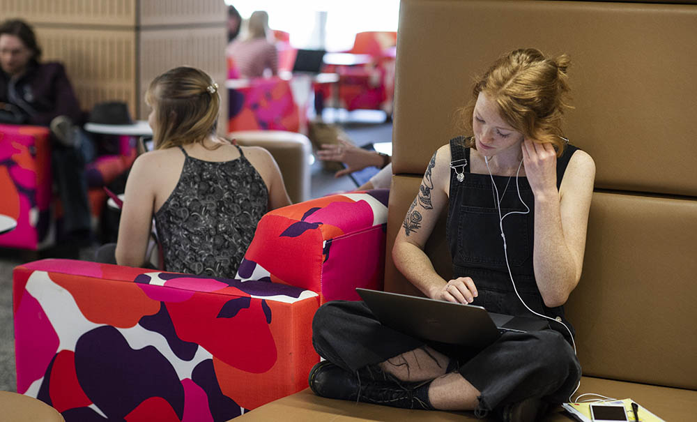 A female student wearing dark overalls sits in the library studying between classes.