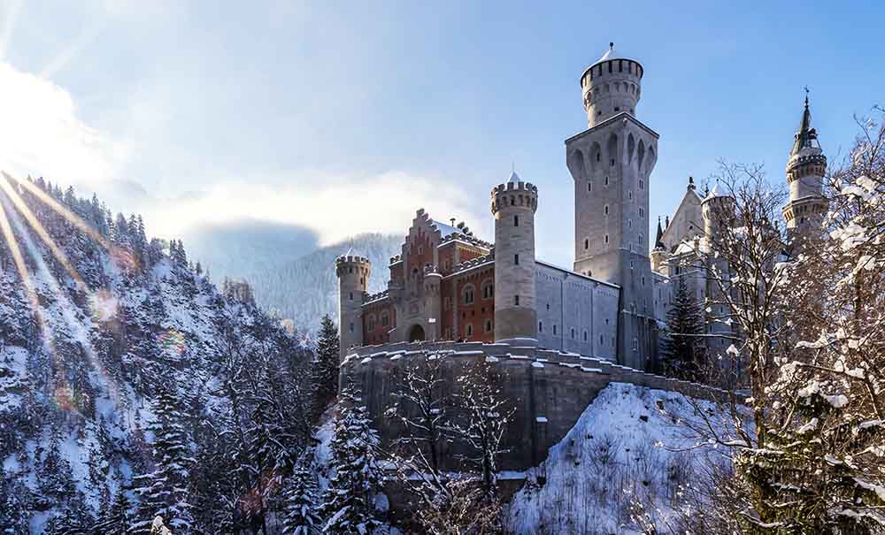 A large stone castle with a tall tower sits on a snowy mountain top.