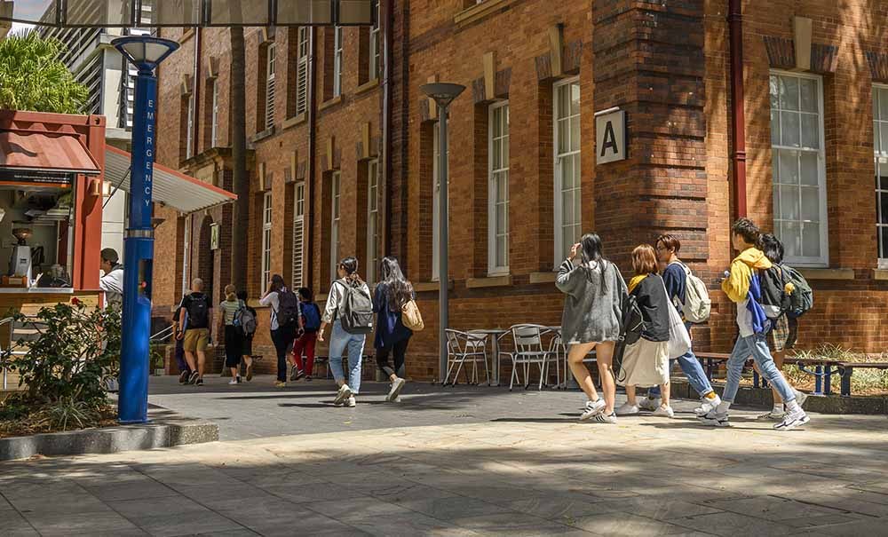 Groups of students walk between two old, deep redbricked buildings. Next to one of the buildings is a coffee cart with students lined up to order.