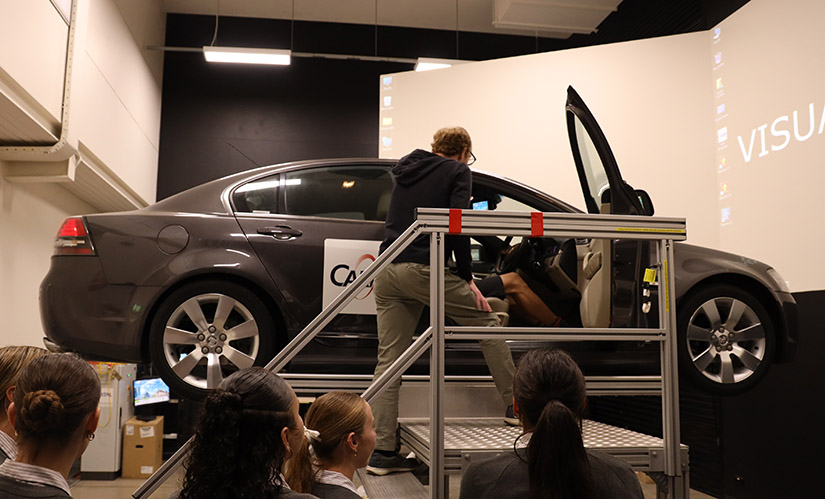 A car is suspended on a mechanical rig. A research participant is in the driver's seat and a male researcher is giving them instructions.
