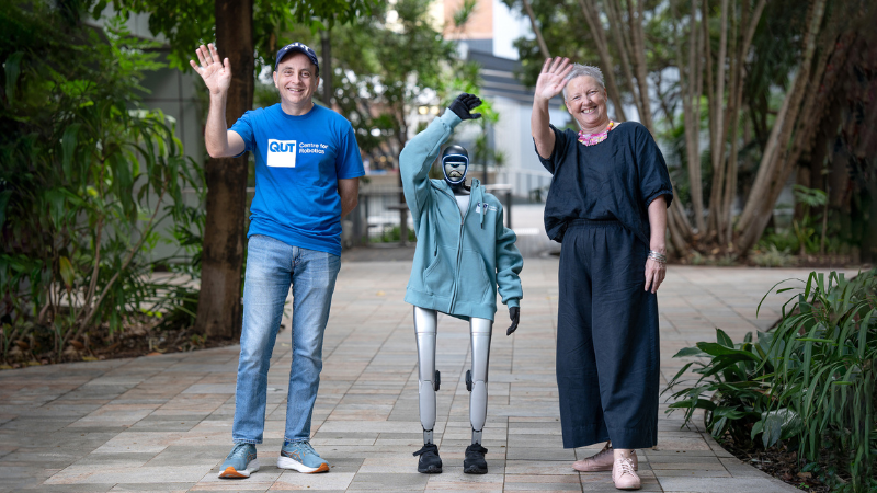Two people stand side beside a small robot. Both the people and the robot are waving their hands.