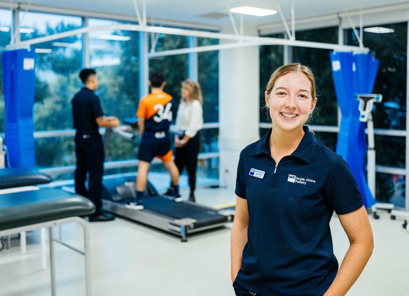 A smiling woman in a clinical uniform stands infront of a treadmill where a patient is doing a gait test.