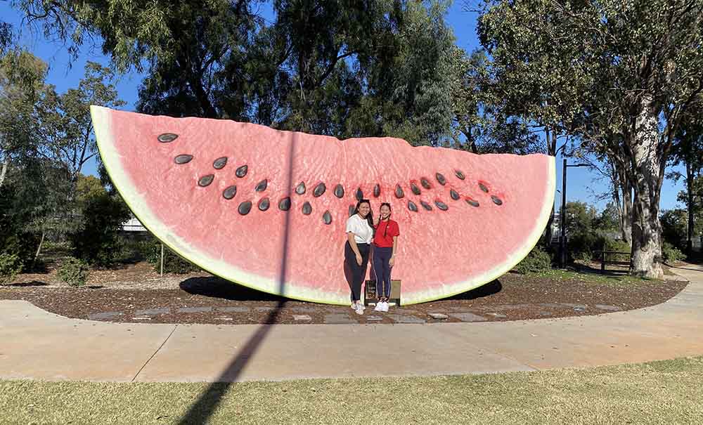 Two female students smile as they stand in front of a giant water melon statue.