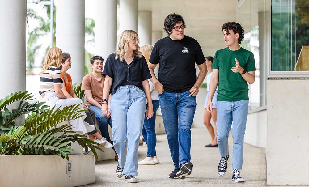 Luke and two friends, all wearing jeans, t-shirts and converse sneakers walking through a university hallway. Luke is telling a story while they walk.