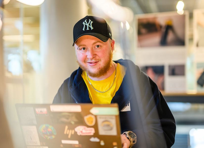 A male student with an eyebrow piercing and beard, wearing a NYC baseball cap and a hoodie sits at a laptop in a bright, modern student space a uni.
