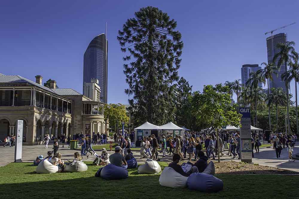 An outdoor campus scene on a warm, winter's day. In the foreground we see people relaxing on bean bags sitting on the grass. In the background busy crowds gather at tents to talk to academics and students.