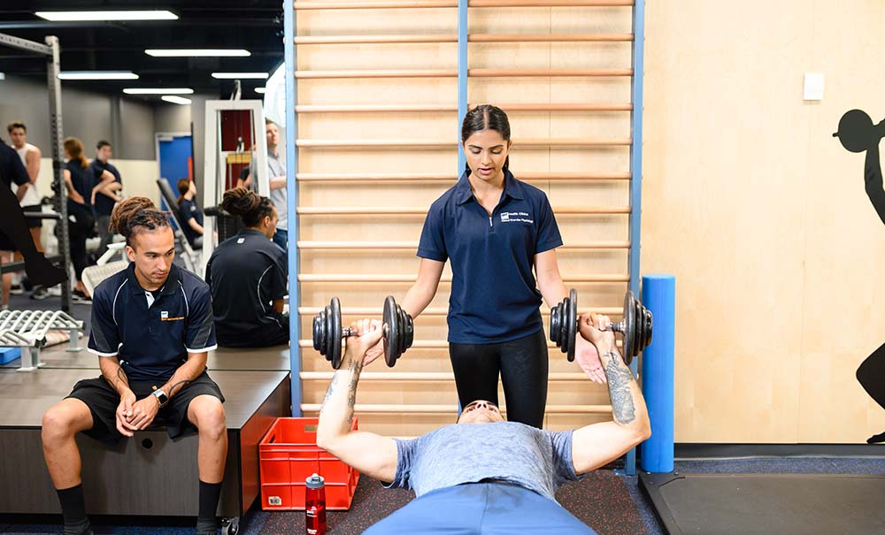 A female student in a QUT sports uniform is standing behind a male client to spot him on the bench press. He is holding large weights and she has her hands underneath the weights ready to catch them.