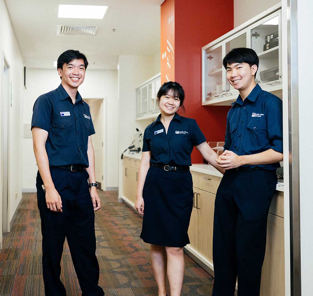 Three smiling optometry students in clinical uniforms stand in a hallway outside of clinical treatment rooms..