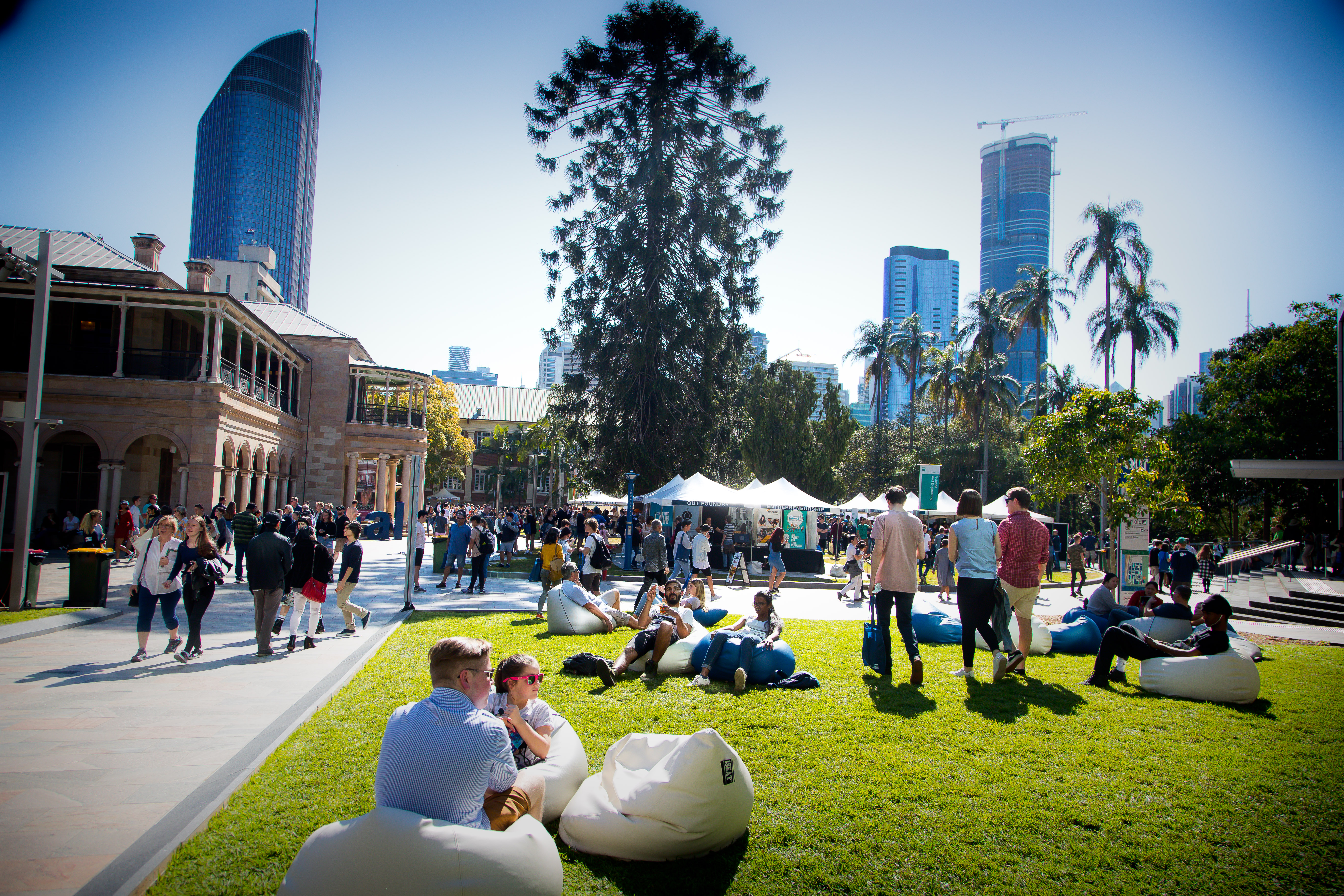 A sunny day on campus, in the foreground people relax on bean bags in the background there are crowded tents.