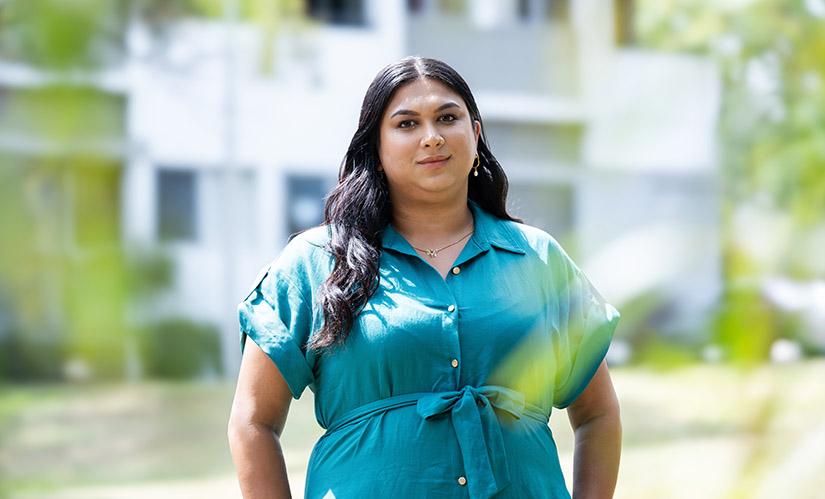 Karyna stands in a sunny park. Behind her is a residential building with multiple apartments.