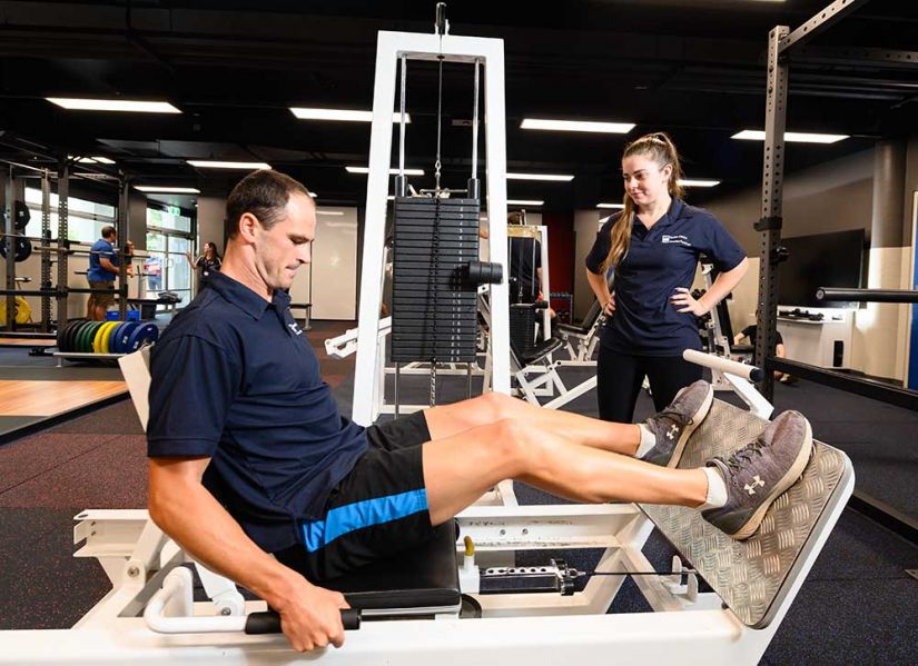 A male athlete uses leg press equipment in a gym while a female exercise science student supervises his form.