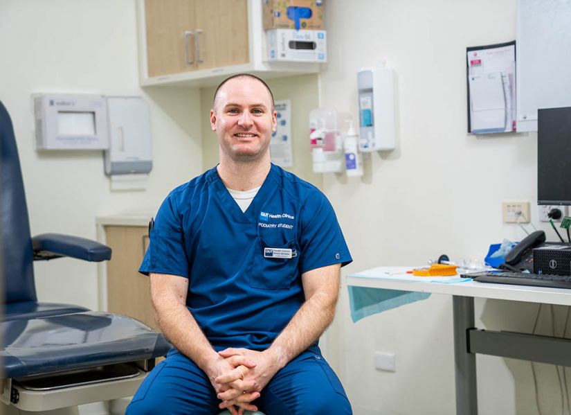 A smiling male student in scrubs sits in a Health clinics consultation room. Next to him is a seat for the patient and on the other side is a desk with computer and equipment.