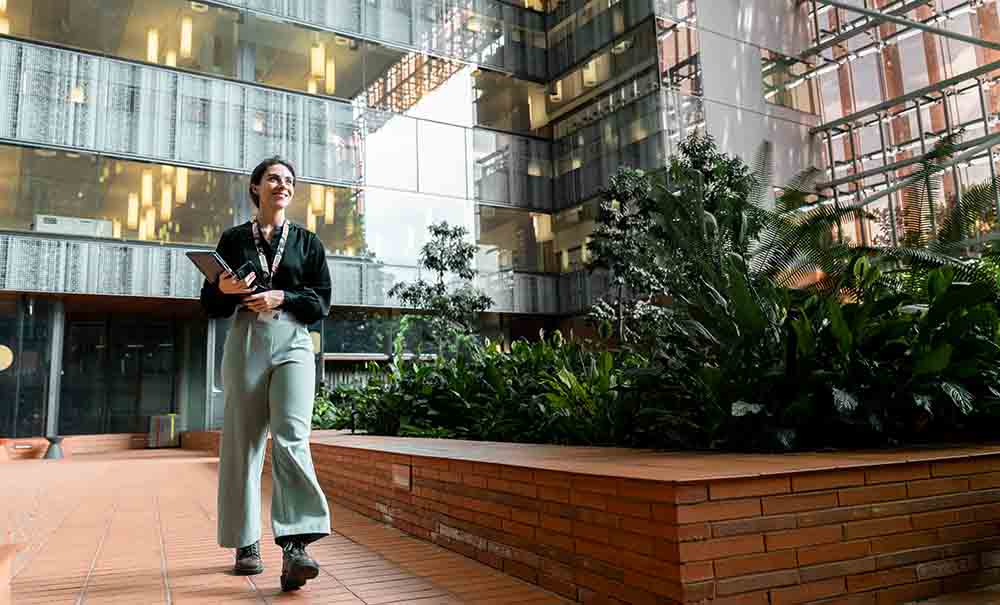 A woman wearing a lanyard, long sleeves and slacks looks up as she walks through a high ceilinged building foyer.