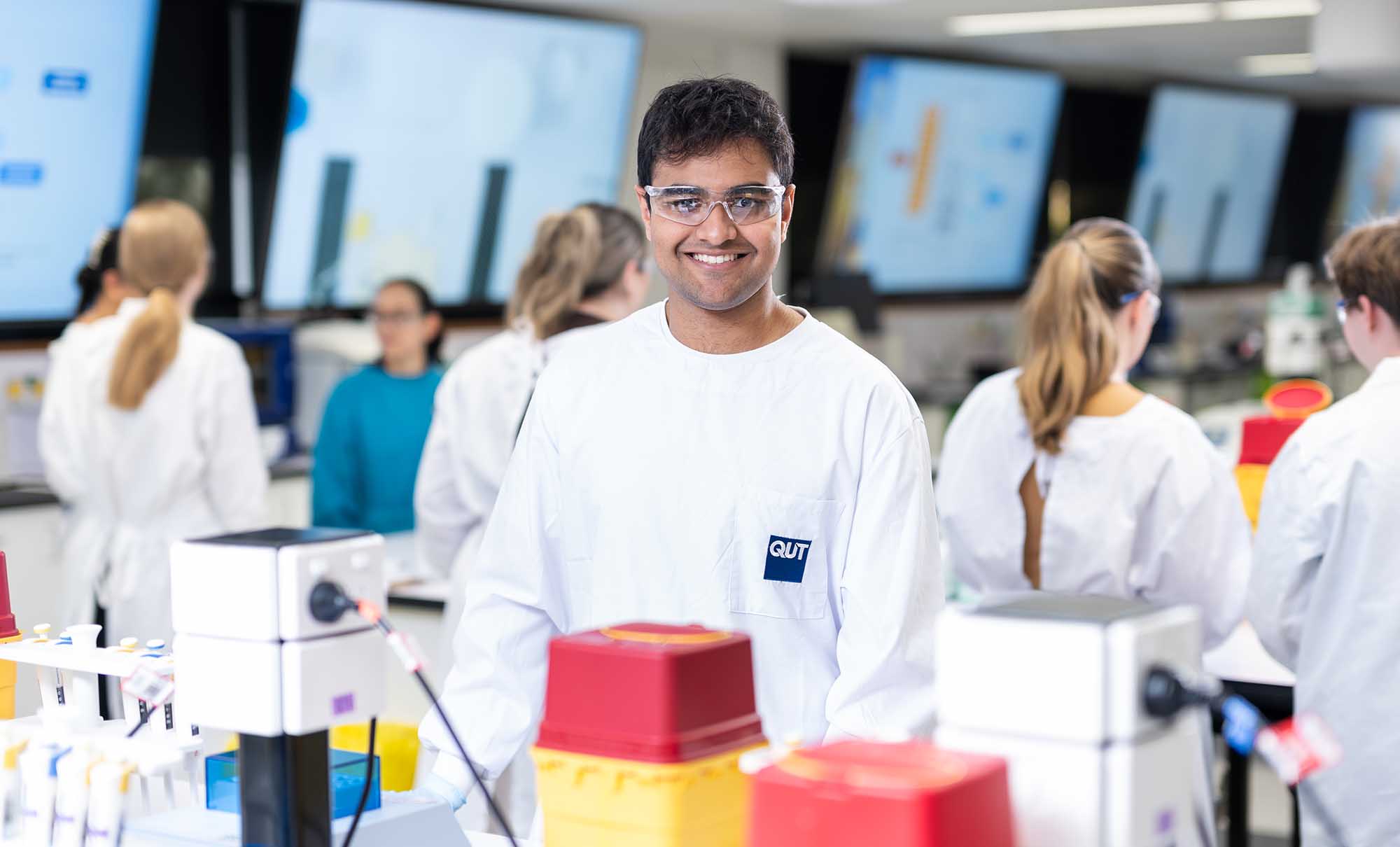A male student stands at a laboratory benchtop, he is wearing a protective gown and glasses.