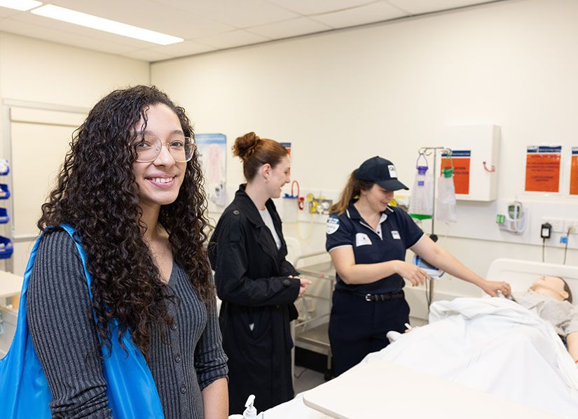A female student with dark, curly hair stands in a classroom that looks like a hospital ward.