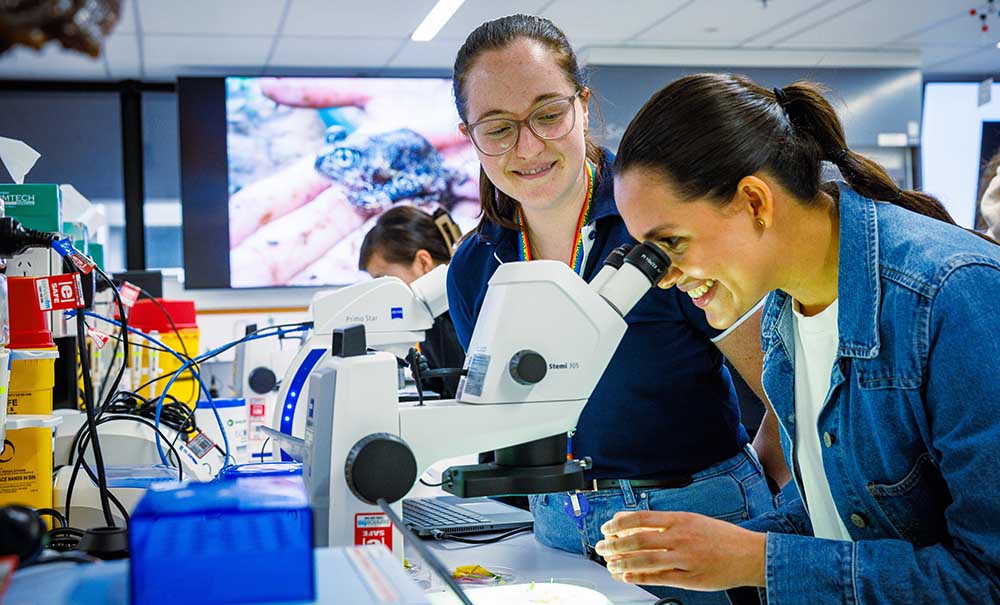 A woman looks down a microscope while a female academic explains what she is looking at.